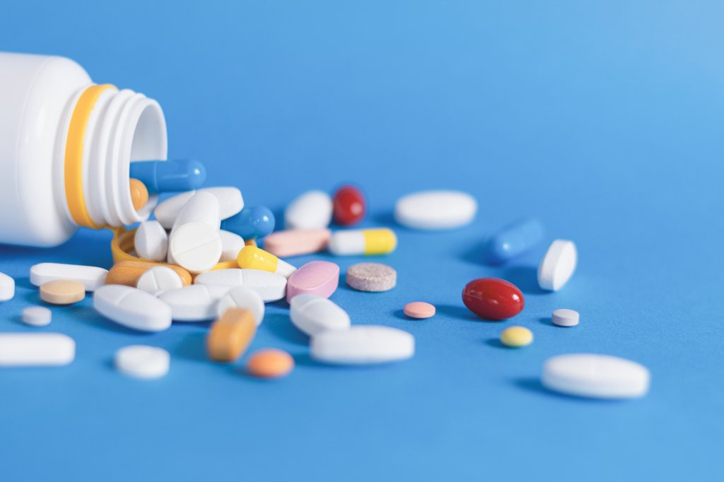 "Close-up stock photo of an open white pill bottle lying on its side on a blue background, with an assortment of different tablets and capsules spilling out — including blue-and-white capsules, white round and oval tablets, and yellow, pink, red, and orange pills of various shapes.